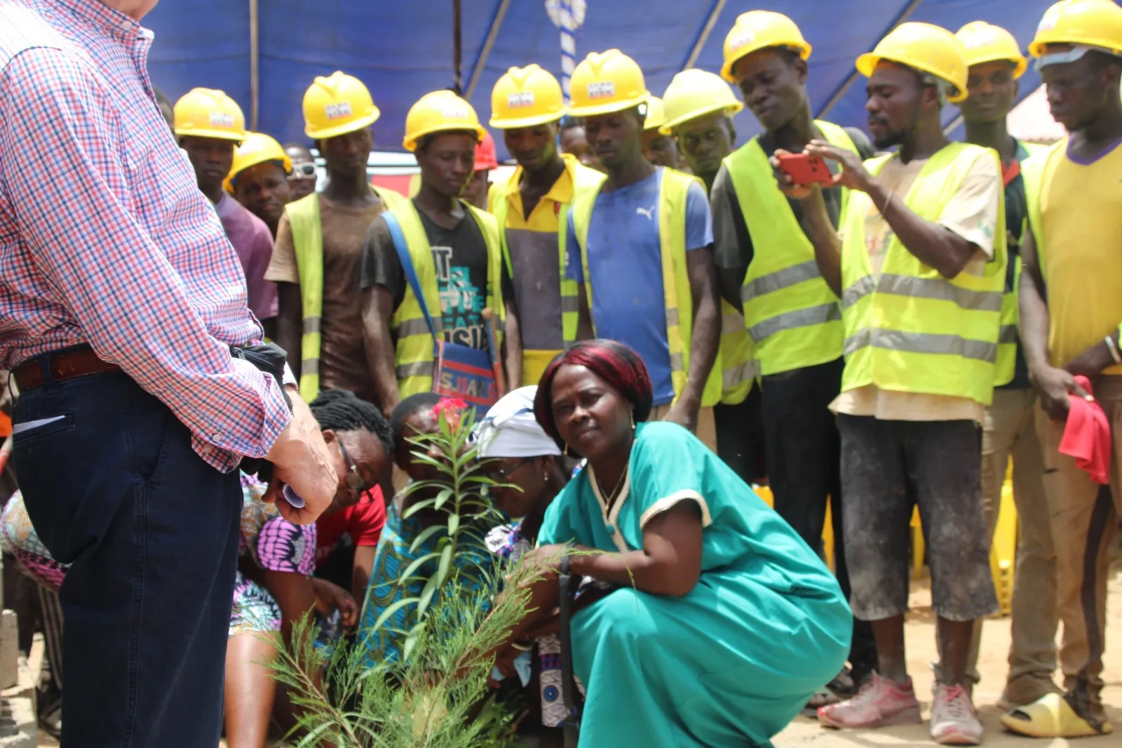 Une femme en tenue verte plante un arbre, entourée des ouvriers du chantier